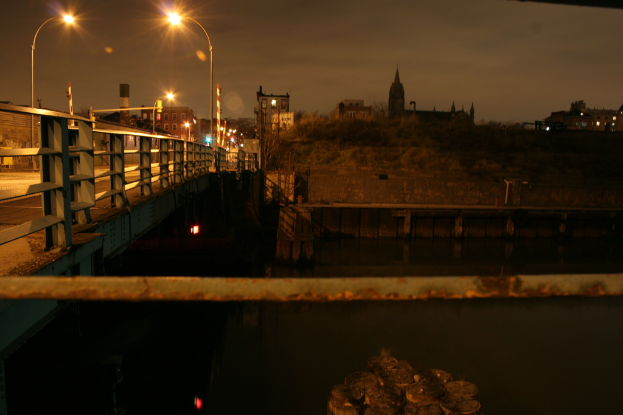 Eine Brücke spannt einen Fluss mit zahlreichen Straßenlaternen darauf und viele Gebäude sind im Hintergrund zu sehen.