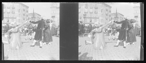 Ein Schwarz-Weiß-Foto einer Gruppe von Menschen in Kostümen, die auf der Straße tanzen, umgeben von Gebäuden, Straßenlaternen und Straßenlaternen unter einem klaren Himmel.