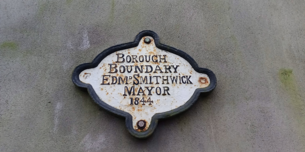 Metallplakette an einer Gebäudeseite, die die Borough-Grenze markiert, mit "Borough Boundary Edm Smithwick Mayor" in fetten weißen Buchstaben auf schwarzem Hintergrund.