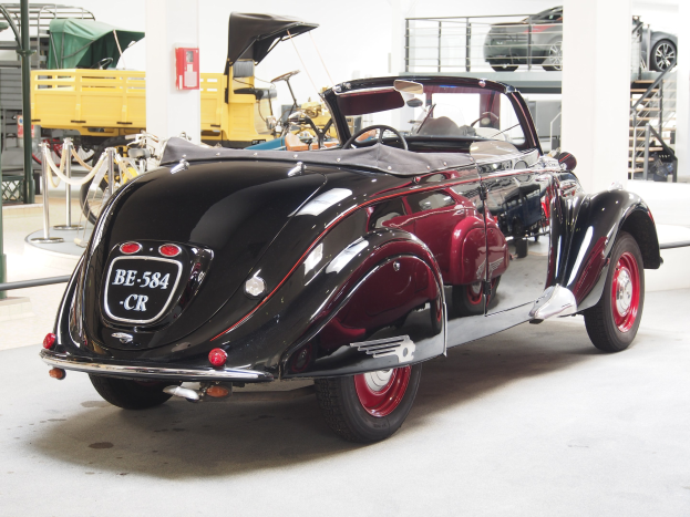 Schwarzes und rotes Auto in einer Garage mit anderen Fahrzeugen, Pfählen, Ketten und einem Feuerlöscher an der Wand im Hintergrund geparkt.