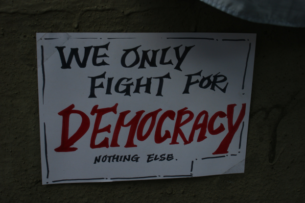 Weißes Papier mit "Wir kämpfen nur für die Demokratie" darauf, an einer Wand befestigt.