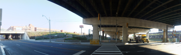 Ein Lastwagen fährt unter einer Brücke auf einer Stadtstraße mit Gebäuden, Straßeninfrastruktur, Fahrzeugen, Grünflächen und einem klaren Himmel.