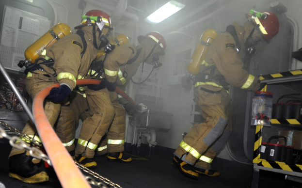Feuerwehrleute in Schutzausrüstung und Helmen arbeiten an einem Feuerwehrauto, mit Equipment-Regalen und Hintergrundrohren im Hintergrund.