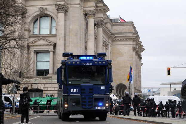 Eine Gruppe von Polizisten steht vor einem großen Gebäude mit Fenstern, Säulen und Bögen, mit Fahrzeugen auf der Straße und einer Person mit einer Kamera auf der linken Seite, unter einem klaren blauen Himmel mit Bäumen, Ampeln und Flaggen im Hintergrund.