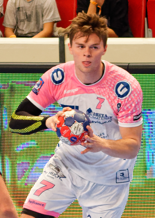 Ein Mann in einem pinken Hemd hält einen Fußball, wobei die Hand einer anderen Person auf der linken Seite des Bildes zu sehen ist und im Hintergrund einige Menschen auf Stühlen sitzen.