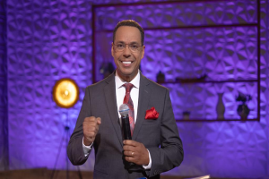 Stephen Colbert, der einen Anzug, Krawatte und Brille trägt, steht auf einer Bühne und hält ein Mikrofon in der Hand, mit einer Wand und verschiedenen Gegenständen dahinter.