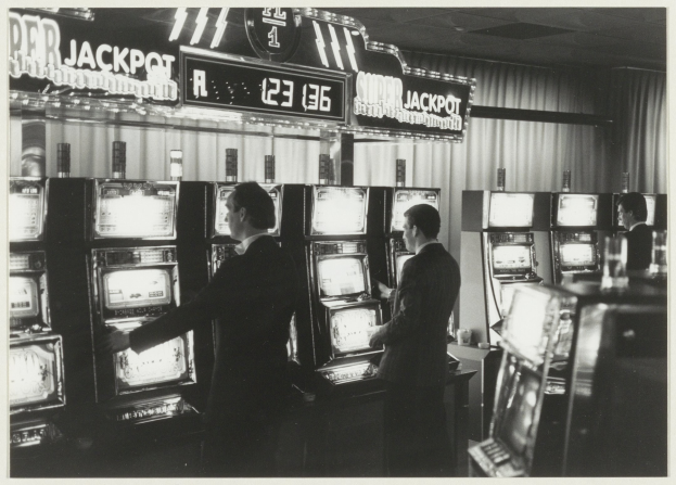 Schwarzes und weißes Foto von zwei Männern, die Spielautomaten in einem Casino spielen, einer hölt ein Glas, mit Vorhängen und einer Texttafel im Hintergrund.