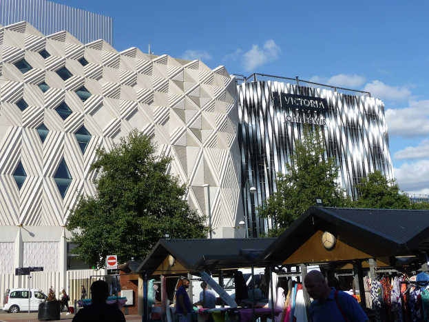 Außenansicht des Victoria and Albert Museums in Birmingham, England, mit Menschen, die gehen, Kleiderständen, Bäumen, Gebäuden und einem bewölkten Himmel.