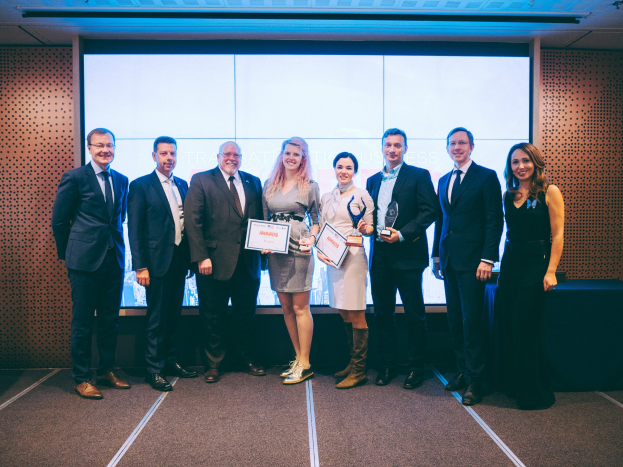 Gruppe von strahlenden Menschen mit Urkunden, die vor einem Bildschirm stehen, mit einem Tisch und einem Objekt an der Wand im Hintergrund, Gewinner der Internationalen Wirtschaftsauszeichnungen 2018.