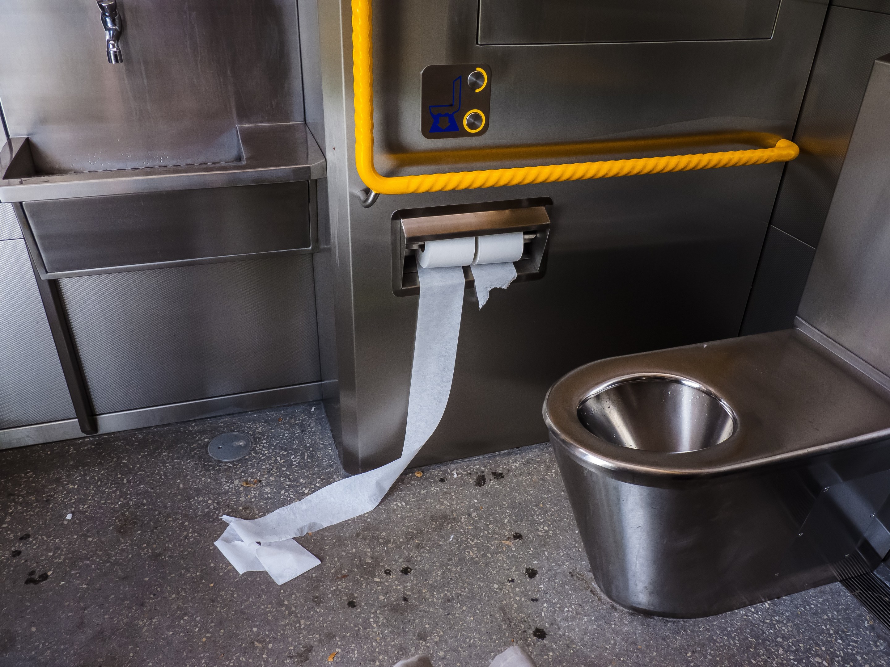 Stahltoilette mit einer Rolle Toilettenpapier daneben, einem Waschbecken mit Wasserhahn und verschiedenen Gegenständen auf dem Boden.