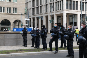 Eine Gruppe von Polizisten in schwarzen Uniformen und Masken steht vor einem Berliner Gebäude mit Glasfenstern und Säulen, einige halten Helme, mit Laternen, Verkehrsampeln, Schildern mit Text, einer Statue und anderen Menschen im Hintergrund.