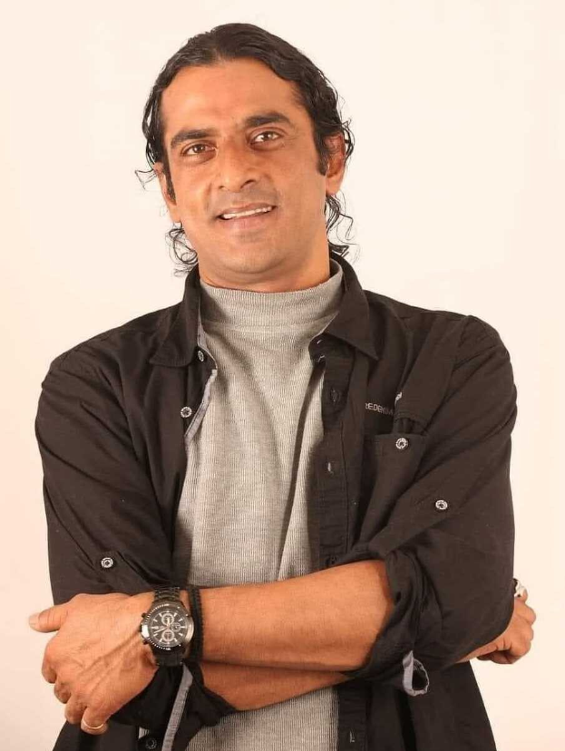 Ein Mann in einem schwarzen und aschfarbenen Kleid mit einer Uhr steht mit verschräckten Armen vor einem weißen Hintergrund und sieht nachdenklich aus.