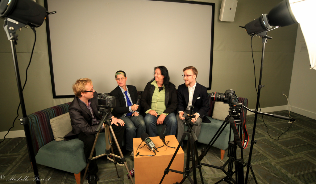 Eine Gruppe von drei Personen sitzt auf einer Couch und gibt ein Interview, wobei eine Person in einem schwarzen Mantel und Hose links, eine Person in einem Mantel und einer blauen Krawatte mit einem Mikrofon in der Mitte und eine Person mit einem Lächeln rechts zu sehen ist. Ein Mikrofonrekorder befindet sich auf dem Tisch in der Mitte, Kameras sind auf der rechten und linken Seite und große Scheinwerfer sind vorhanden, mit einem Projektionsschirm im Hintergrund.