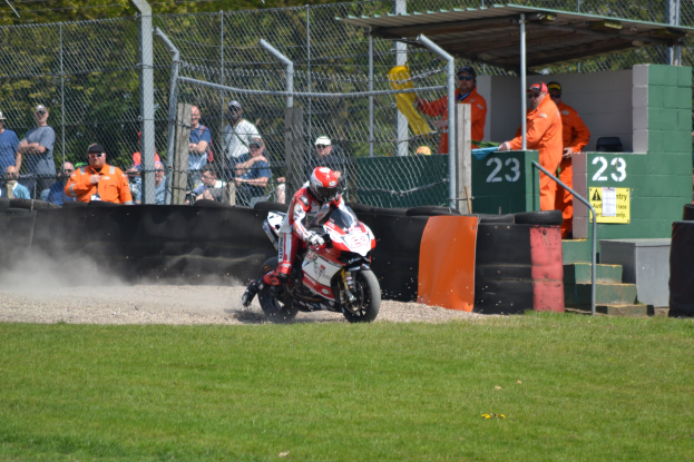 Ein Motorradfahrer mit Helm und Handschuhen fährt über ein grasbewachsenes Feld, mit Zuschauern in Mützen und Schutzbrillen, einem Zaun, einer Treppe mit Geländer und einem nummerierten Schuppen im Hintergrund.
