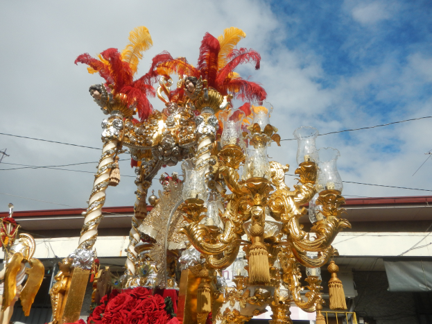 Ein großer goldener und roter Festwagen, der mit Blumen und anderen Dekorationsgegenständen geschmückt ist, bei einem Karnevalsumzug mit einem Gebäude, Strommasten mit Drähten und einem bewölkten Himmel im Hintergrund.