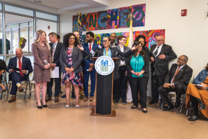 Gruppe von Menschen in einem Raum mit einem Podium, einem Mikrofon und einer Tafel mit der Aufschrift "Sancho-Stadtrat tritt dem Stadtrat bei", umgeben von Fahnen, Stühlen und einem Feuerlöscher.