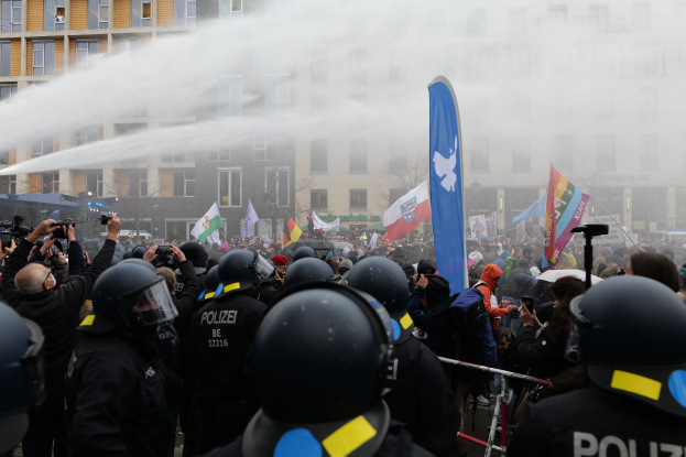 Eine Gruppe von Polizisten steht vor einer Menge, einige halten Kameras und tragen Helme, während ein Wasserwerfer Wasser versprüht; Gebäude, Bäume und ein klarer blauer Himmel sind im Hintergrund zu sehen.