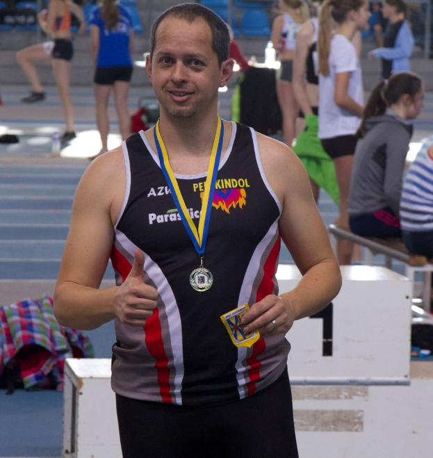 Ein Mann in schwarzem Trikot und Medaille steht, während einige Menschen im Hintergrund trainieren.