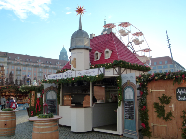 Kleines Gebäude mit rotem Dach und einem Riesenrad davor, umgeben von Festdekorationen und Menschen, mit Gebäuden, Bäumen und bewölktem Himmel im Hintergrund bei einem Oktoberfest-Event.