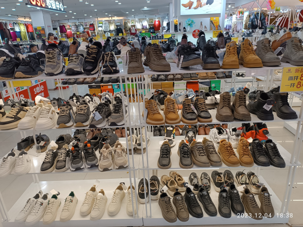 Ein Schuhgeschäfts-Innenraum mit verschiedenen Schuhen auf Gestellen, Preisetiketten sichtbar, Menschen im Hintergrund, Kleider an Bügeln, Deckenlampen, ein Bildschirm und Informationsschilder.