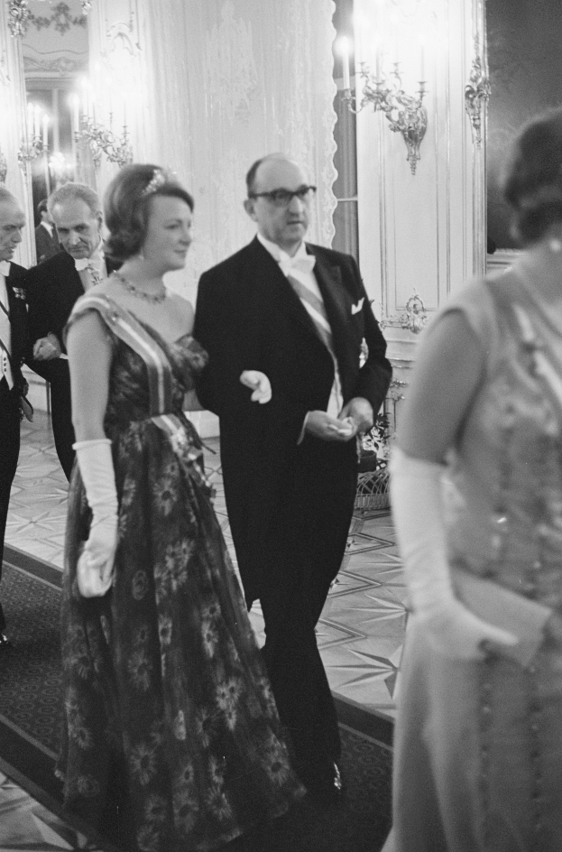 Ein Mann in einem schwarzen Blazer, weißem Hemd und schwarzen Hosen sowie eine Frau in einem eleganten Kleid gehen einen Flur entlang, umgeben von einer Gruppe von Menschen, die auf dem Boden stehen, während ein paar Wandlampen eine gemütliche Atmosphäre schaffen.
