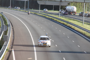 Ein Auto auf einer Autobahn mit einem stationären Polizeiwagen am Straßenrand, andere Fahrzeuge und Bäume, Geländer, Pfosten und Bretter im Hintergrund.