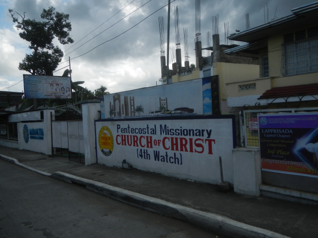 Die Pfingstmissionarische Kirche Christi 14. Wache steht mit einer Straße im Vordergrund, einer Wand mit Bannern und Text, Gebäuden, Bäumen, Masten, Drähten und einem bewölkten Himmel im Hintergrund.