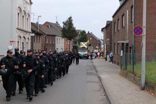 Eine Gruppe von Polizisten in schwarzen Uniformen und Helmen geht eine Straße entlang, die von Gebäuden gesäumt ist, ein Schild an einem Pfahl und Bäume im Hintergrund.