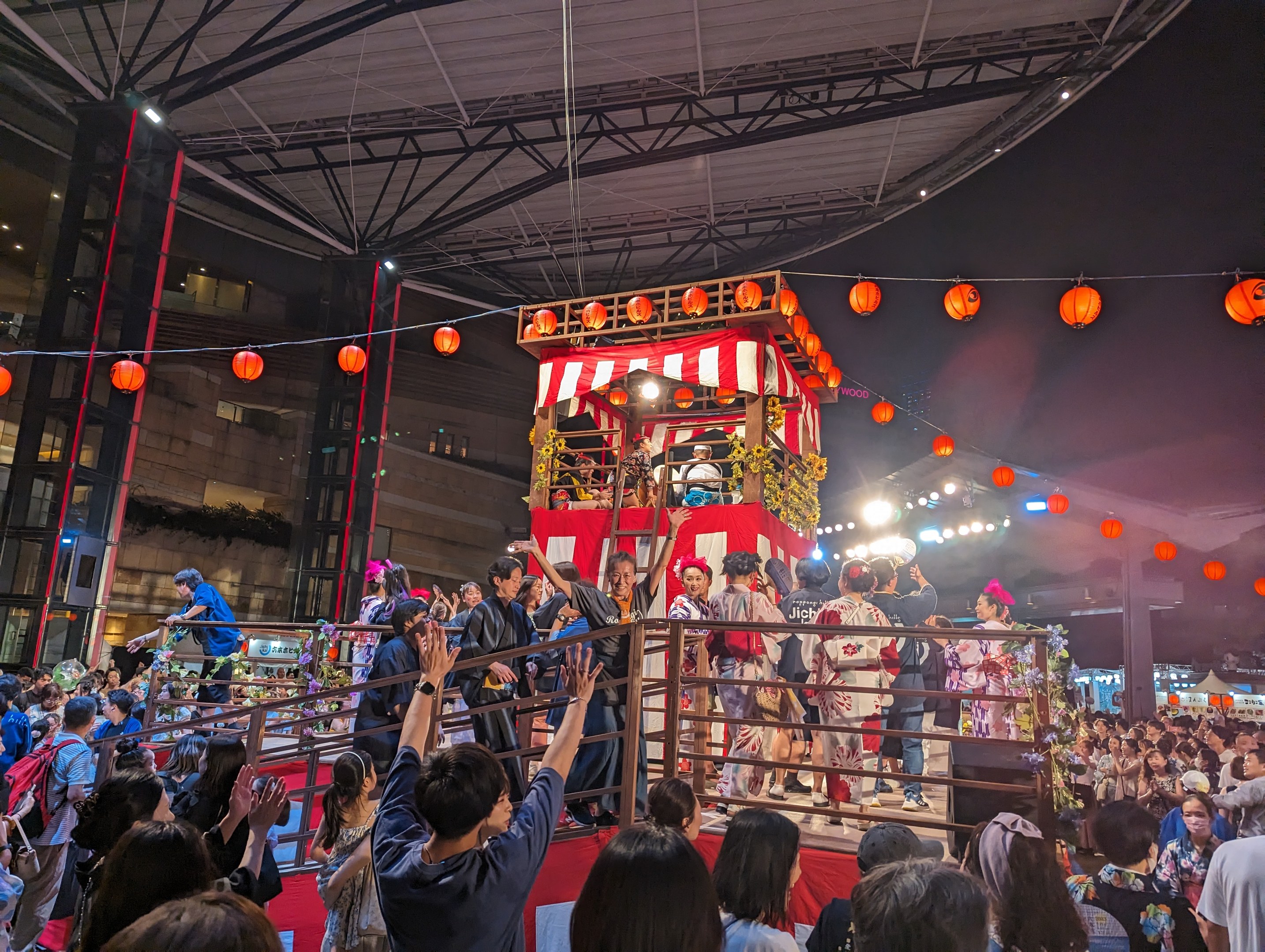 Eine Menschenmenge auf einer beleuchteten Bühne mit Laternen, einige halten Kameras, umgeben von einem Geländer, mit Bäumen und einem klaren blauen Himmel im Hintergrund bei Nacht, wahrscheinlich bei einem japanischen Festival.