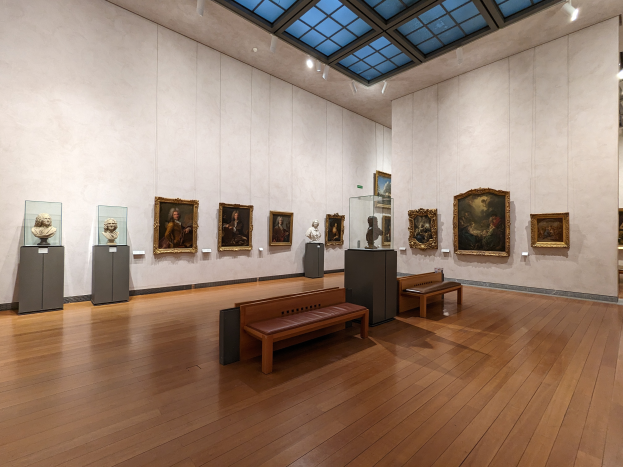 Ein geräumiger Raum im Metropolitan Museum of Art in New York City, der zahlreiche Gemälde an den Wänden, Bänke auf dem Boden, Statuen auf Ständern und Lampen an der Decke zeigt.