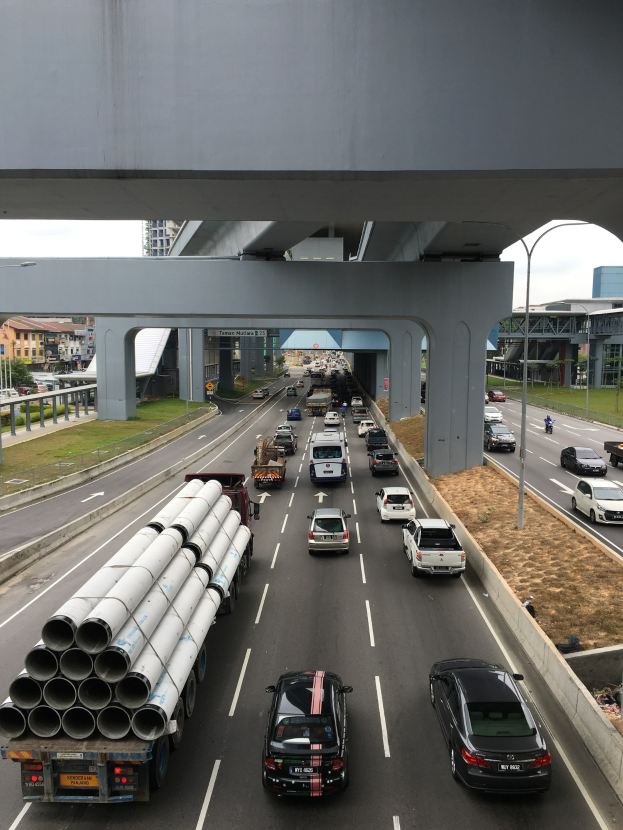 Starker Verkehr auf einer Autobahn unter einer Brücke, mit Straßenlaternen, Gras, Gebäuden, Bäumen und dem Himmel im Hintergrund.