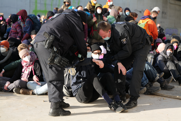 Eine Gruppe von Menschen, einige mit Mützen und Masken, sitzt auf dem Boden, während zwei Polizeibeamte in der Nähe stehen, in einer städtischen Umgebung, mit einer Wand voller Graffiti im Hintergrund.