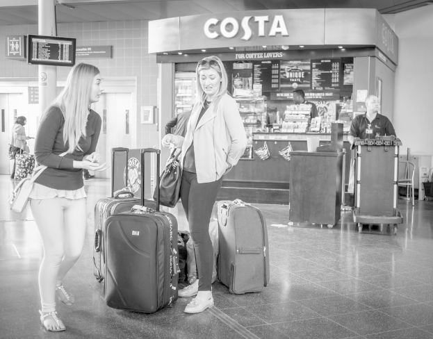 Zwei Frauen mit Gepäck vor einem Kaffeehaus in einer Art Flughafen.