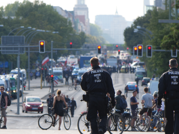 Zwei Polizisten neben einer Gruppe von Radfahrern auf einer Straße mit Fahrzeugen, Verkehrszeichen, Bäumen, Gebäuden und einem klaren blauen Himmel.
