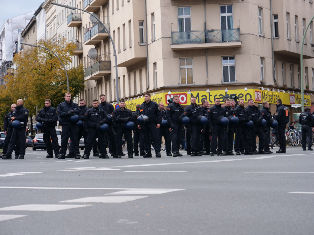 Eine Gruppe von Polizisten in schwarzen Uniformen mit blauen Helmen steht in einer Straße, die von Laternenmasten, Bäumen und Gebäuden mit Glasfenstern und Balkonen gesäumt ist, unter einem klaren blauen Himmel.