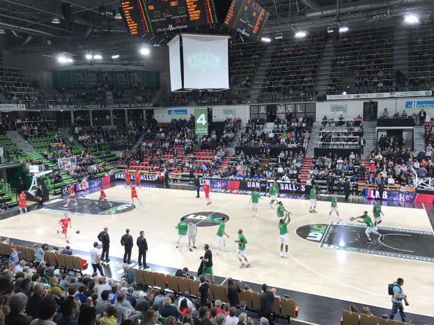 Eine Gruppe von Menschen, die Basketball in einer vollen Halle spielen, mit Zuschauern auf den Tribünen und beleuchtetem Spielfeld.