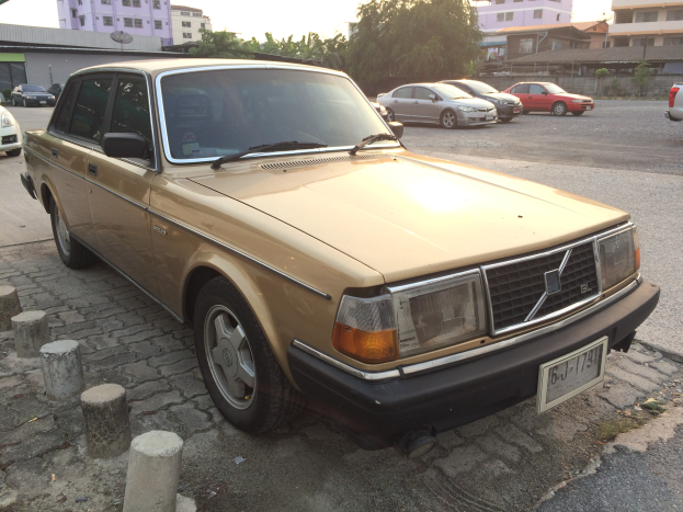 Ein goldfarbener Wagen am Straßenrand mit Gebäuden, Bäumen und einem klaren blauen Himmel.