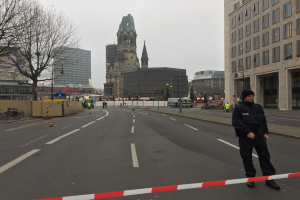 Ein Polizist in schwarzer Uniform und Mütze steht auf einer Straße in Berlin umgeben von Verkehrskegeln, Fahrrädern, Laternenpfählen, Bäumen und Gebäuden, mit einem Kirchturm und blauem Himmel im Hintergrund.