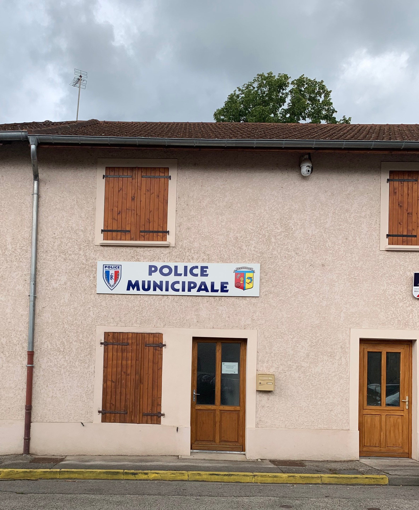 Gebäude der Polizei mit einem Schild, Fenstern, Türen, einer Namensplakette, einem Rohr, einem Baum und einem bewölkten Himmel im Hintergrund.