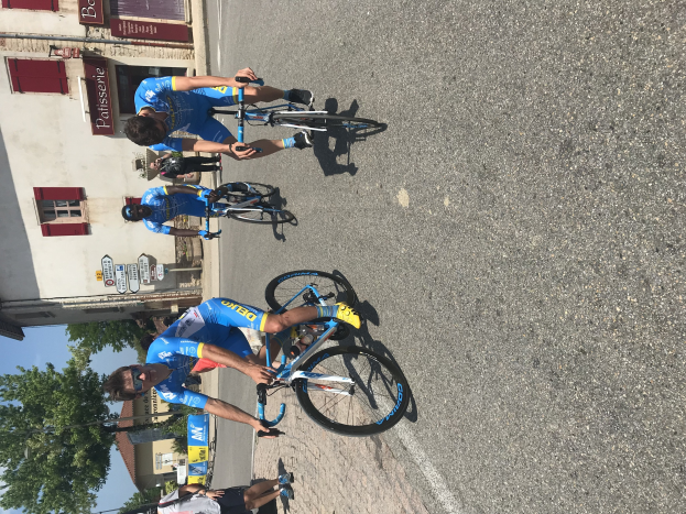 Eine Gruppe von Radfahrern fährt eine Straße entlang an einem Gebäude mit vernagelten Fenstern und Text vorbei, Bäumen, einem klaren Himmel und Zuschauern auf der Straße darunter.