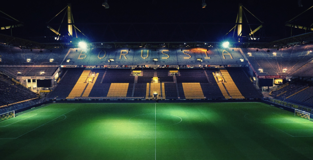 Ein Fußballfeld bei Nacht, erhellt von hellen Stadionlichtern, mit ein paar Menschen auf den Tribünen und grünem Rasen auf dem Boden.