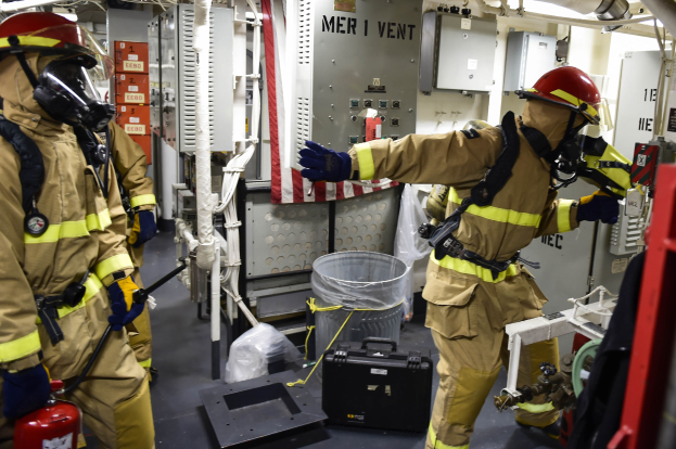 Drei Feuerwehrleute in Schutzausrüstung stehen neben einem Feuerlöscher in einem unordentlichen Raum mit Rohren, Lampen und Equipment im Hintergrund.