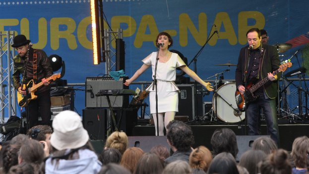 Eine Buhnenbühne mit vier Performern: eine Frau in einem weißen Kleid, die in der Mitte singt, zwei Männer, die links und rechts Gitarren spielen, eine Menge vorne und Lautsprecher im Hintergrund.