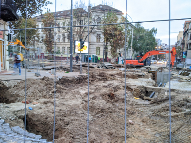 Baustelle mit einem ausgehobenen Bereich, der von einem Metallzaun umgeben ist, angrenzende Straße mit Fahrzeugen und Fußgängern, Verkehrsampeln, Schilder, Bäume, Gebäude und klarer Himmel.