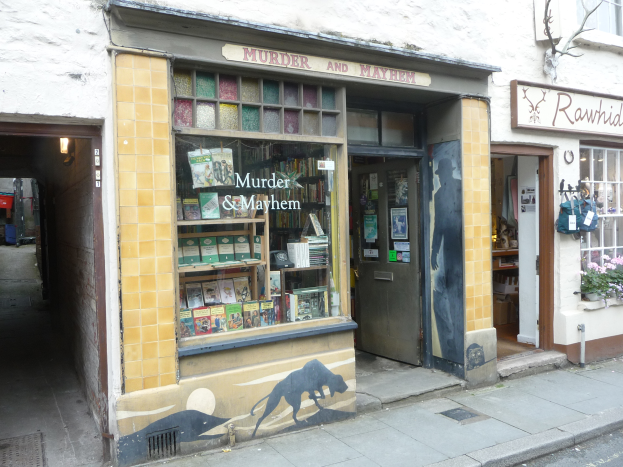 Ein Geschäft namens 'Mord und Totschlag' mit Regalen voller Bücher, verschiedener Gegenstände, Texttafeln und Pflanzen mit Blumen, einem Baum und einer Straße im Vordergrund.