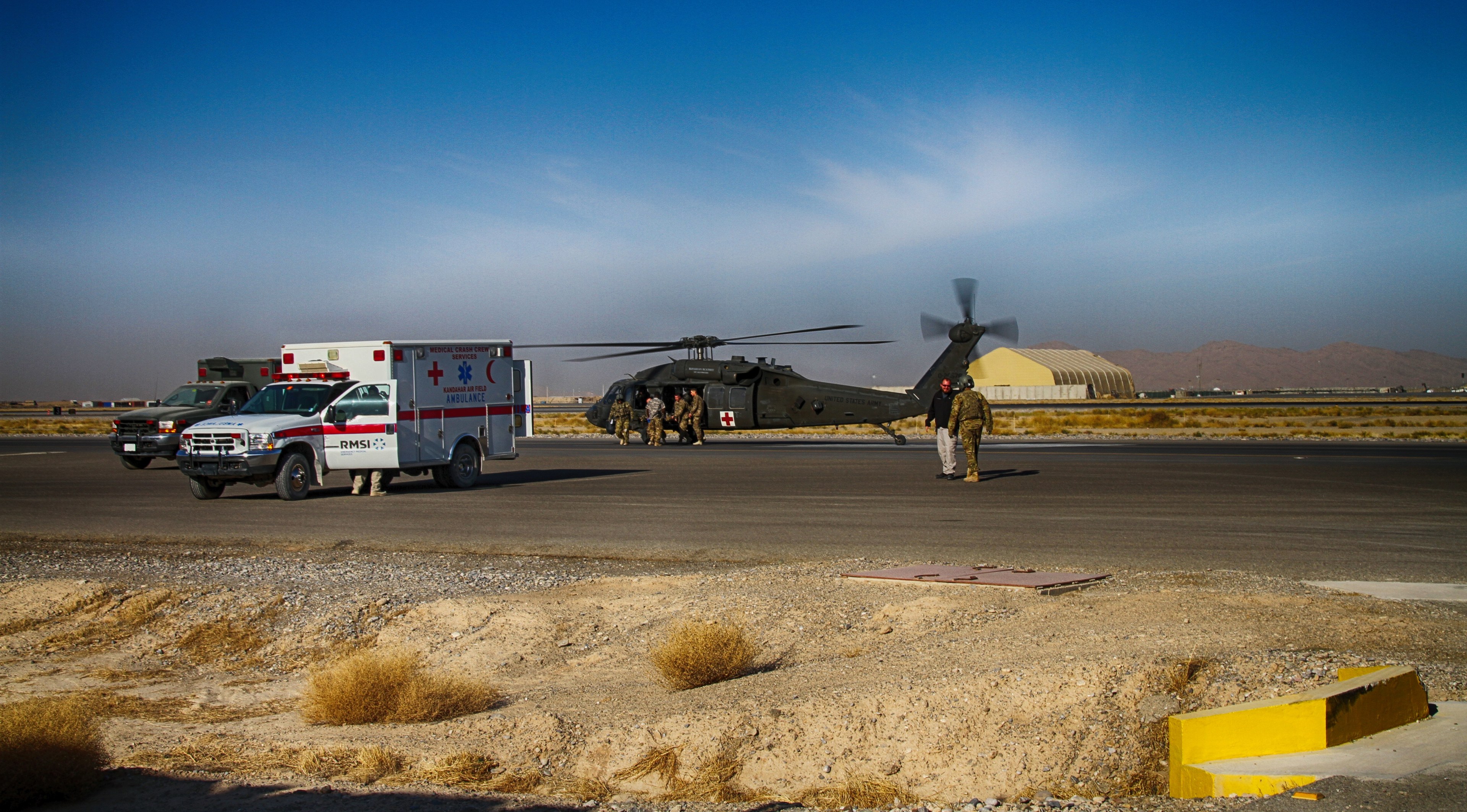 Ein Hubschrauber neben einem Rettungswagen auf einer grasbewachsenen Straße geparkt, mit Menschen in der Nähe und Bergen im Hintergrund unter einem klaren blauen Himmel.