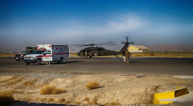 Ein Hubschrauber neben einem Rettungswagen auf einer grasbewachsenen Straße geparkt, mit Menschen in der Nähe und Bergen im Hintergrund unter einem klaren blauen Himmel.