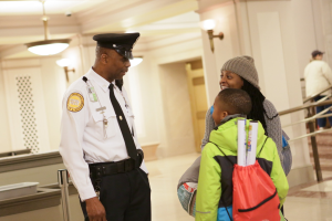 Ein Polizeibeamter spricht mit einer Frau in einer Mütze und einem Kind an einem Flughafen, mit anderen Reisenden im Hintergrund.