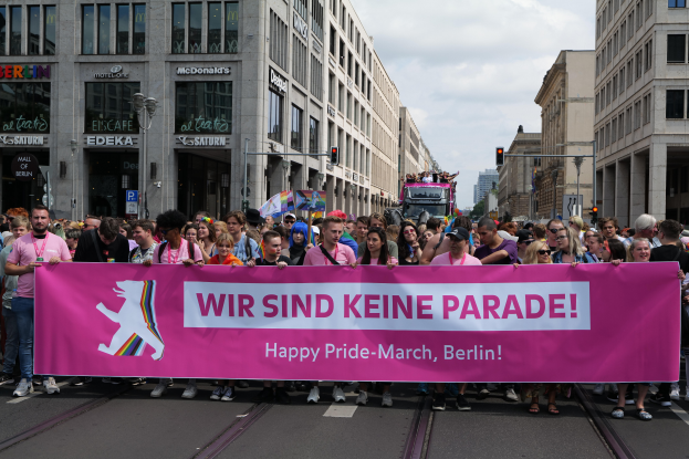 Eine Gruppe von Menschen marschiert auf einer Straße in Berlin, Deutschland, mit einer pinken "Happy Pride March"-Schleife, mit Gebäuden, Laternenmästen und Verkehrszeichen an der Straße entlang unter einem bewölktem Himmel.