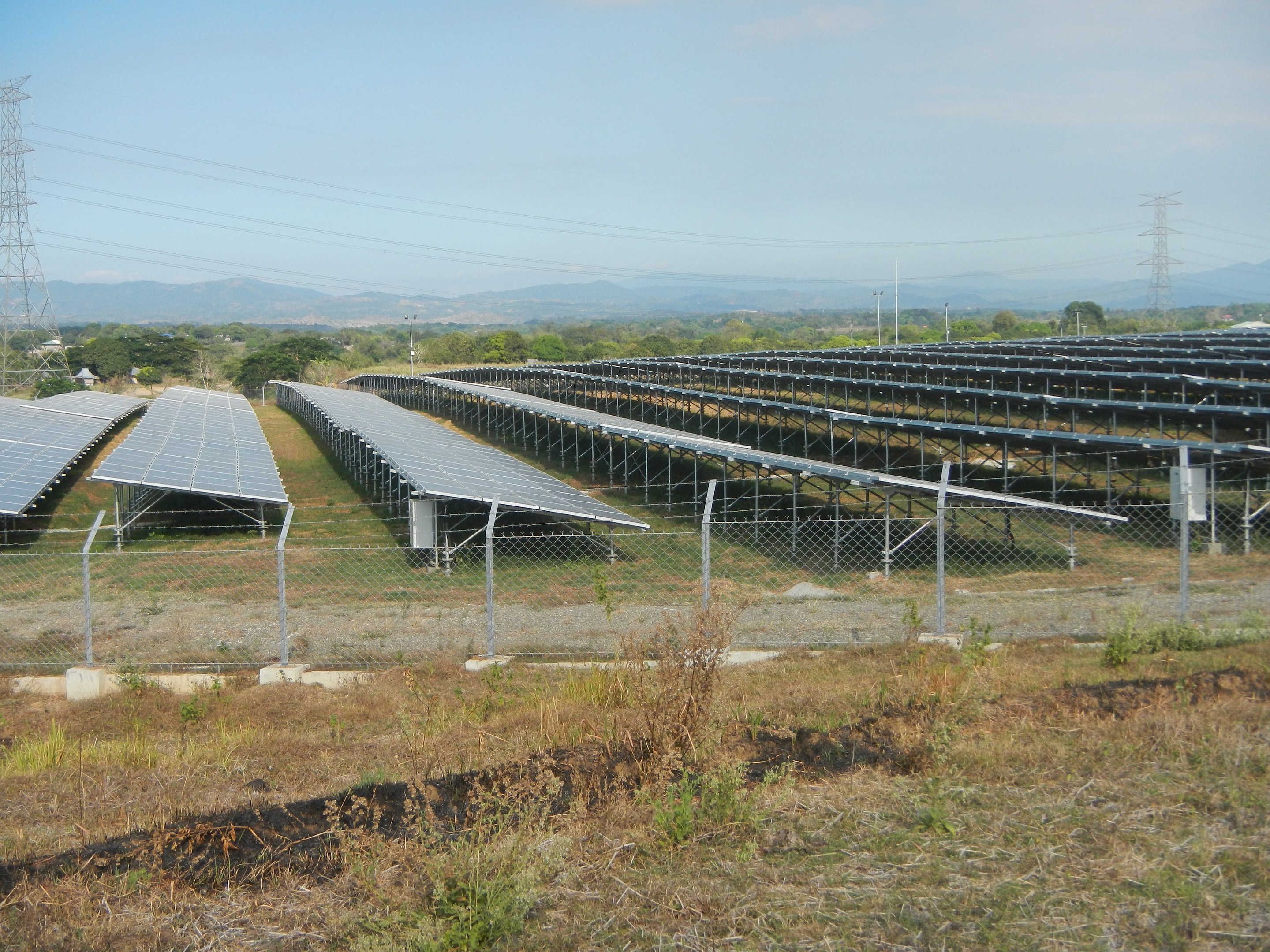Ein Solarpark mit einem Zaun drumherum, davor Gras und Pflanzen, Bäume, Übertragungsmasten mit Drähten und Hügel im Hintergrund unter einem klaren blauen Himmel.